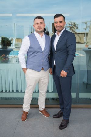 Two people in formal attire pose together at an outdoor venue with glass windows and palm trees in background.