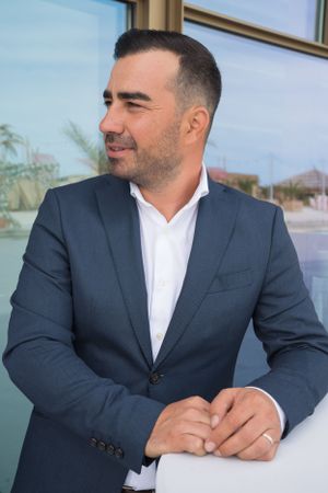 Person in navy suit and white shirt sits at outdoor venue with reflective windows.