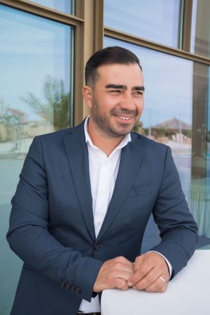 Person in business attire smiles while seated at modern outdoor venue with glass windows.