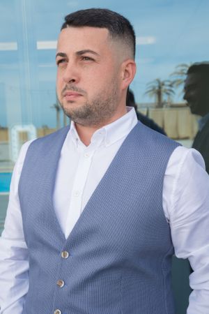 Person wearing white shirt and gray vest poses outdoors against glass building facade.