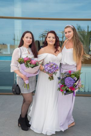 Wedding party members hold colorful bouquets of purple and pink flowers outdoors.