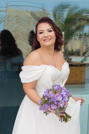 Bride in off-shoulder white wedding dress holds purple orchid bouquet against tropical palm background.