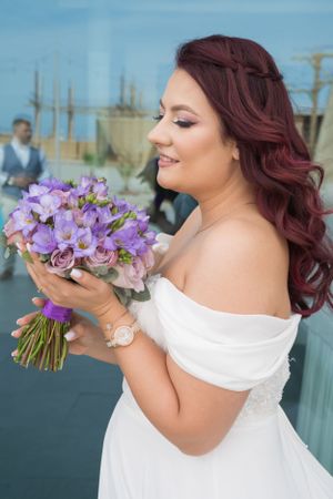 Bride with burgundy hair admires purple orchid bouquet during outdoor wedding photoshoot.