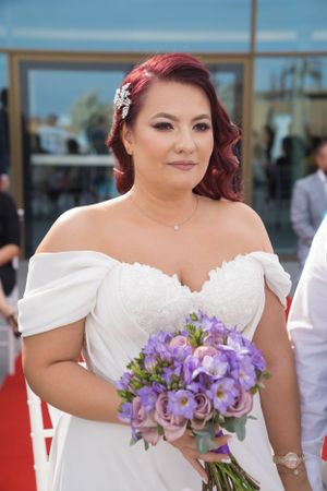 Bride in white off-shoulder dress holds purple floral bouquet.