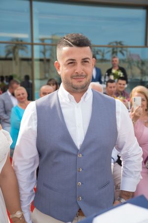 Groom wearing light gray vest and white shirt smiles during outdoor ceremony.