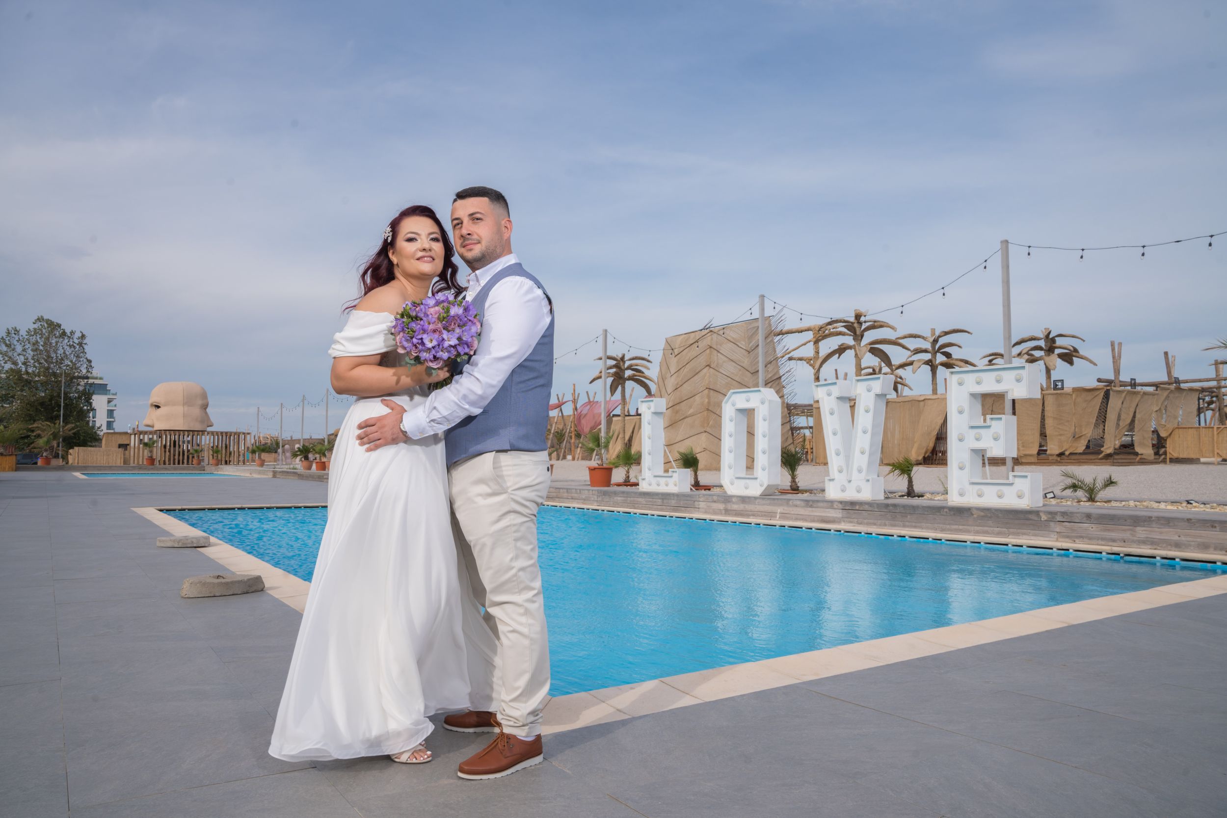 Couple poses for wedding photos at outdoor venue with swimming pool and palm trees.