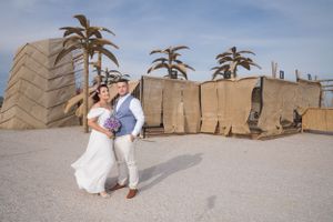 A couple poses against a desert backdrop with palm trees and sandy terrain.