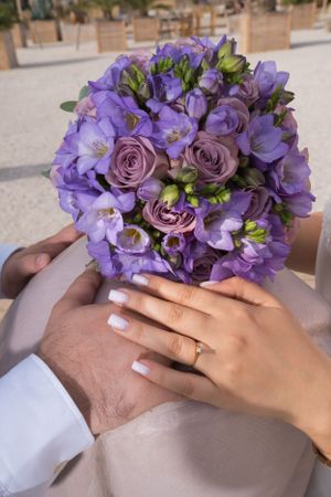 Purple floral bouquet with roses and delicate blooms held by hands with a wedding ring.