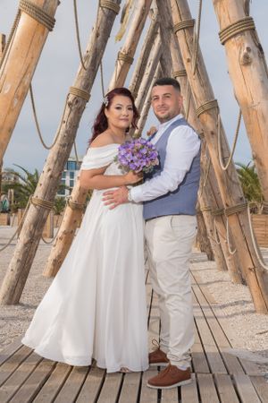 Couple poses romantically on wooden boardwalk with bamboo poles and purple flowers at beach wedding.