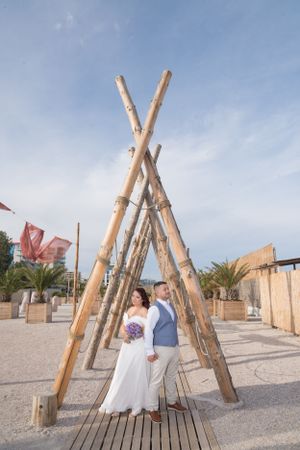 Wedding couple stands between rustic bamboo poles on beachfront wooden deck.