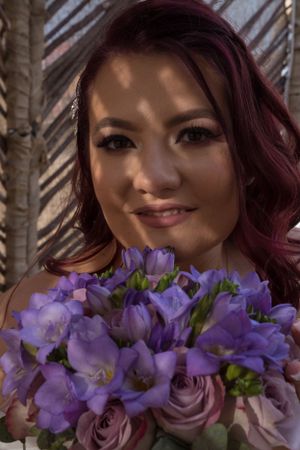 Purple and rose wedding bouquet held near bride's face in artistic portrait.