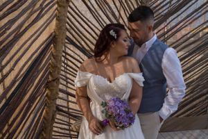 Romantic wedding couple embraces against bamboo wall with purple bouquet.