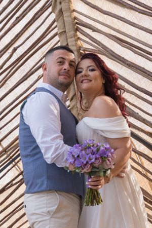 A romantic wedding couple poses against bamboo backdrop holding a purple bouquet.