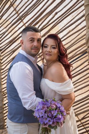 Newlyweds share intimate moment with purple floral bouquet against striped background.