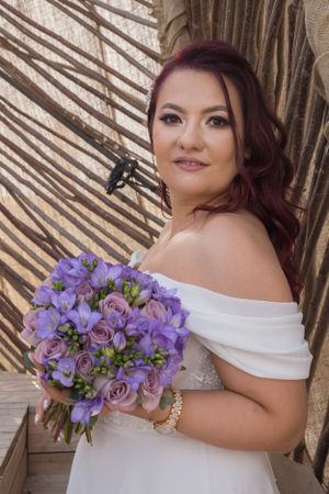Bride in off-shoulder dress holds purple bouquet against natural bamboo backdrop.