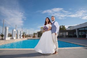 A couple in white attire poses for romantic wedding photos by a modern swimming pool on a sunny day.