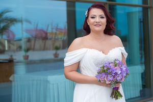 Bride in off-shoulder white gown holds purple floral bouquet against blue glass building.