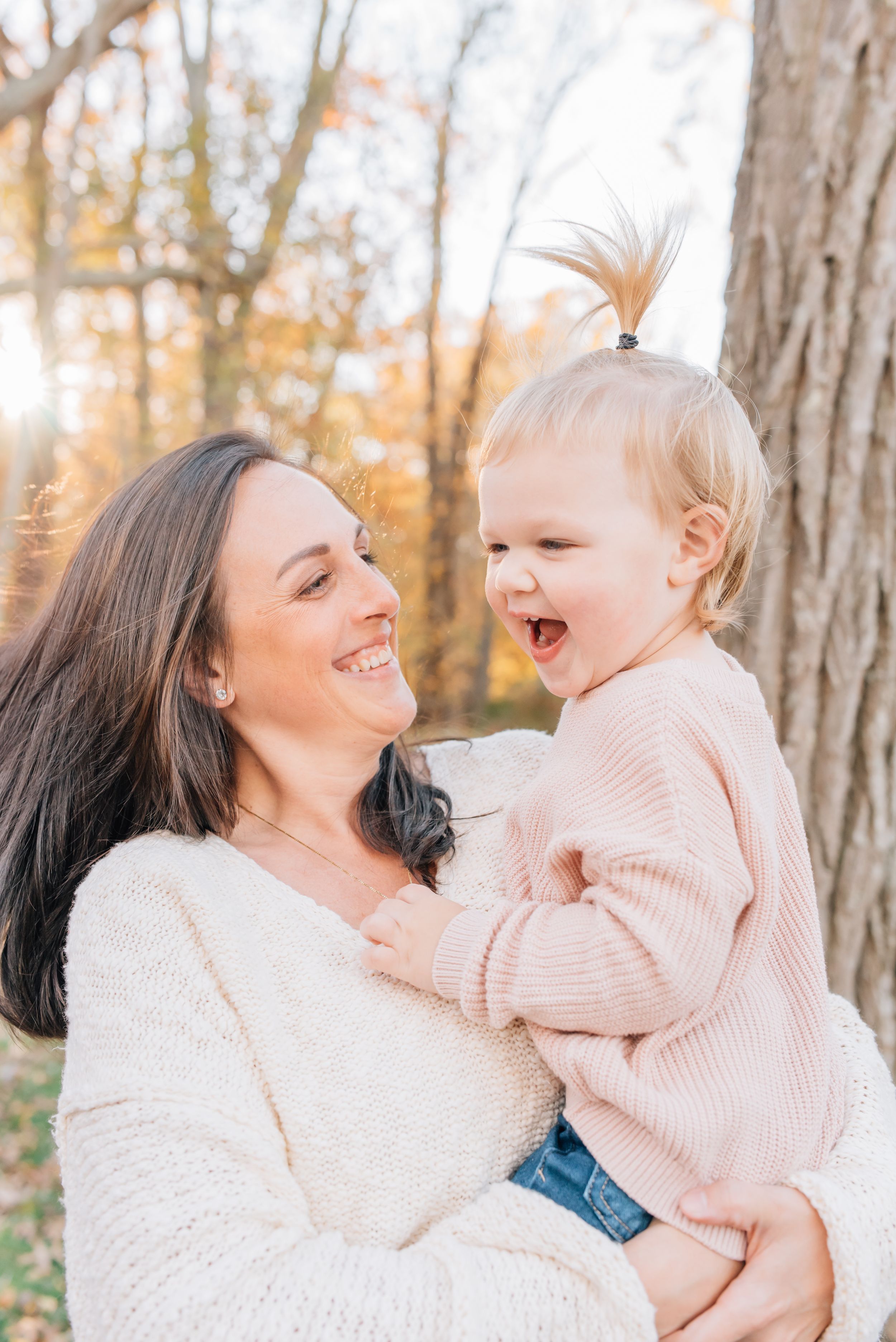 Mother holds her little girl at the park with fall leaves and a sun shining behind them.