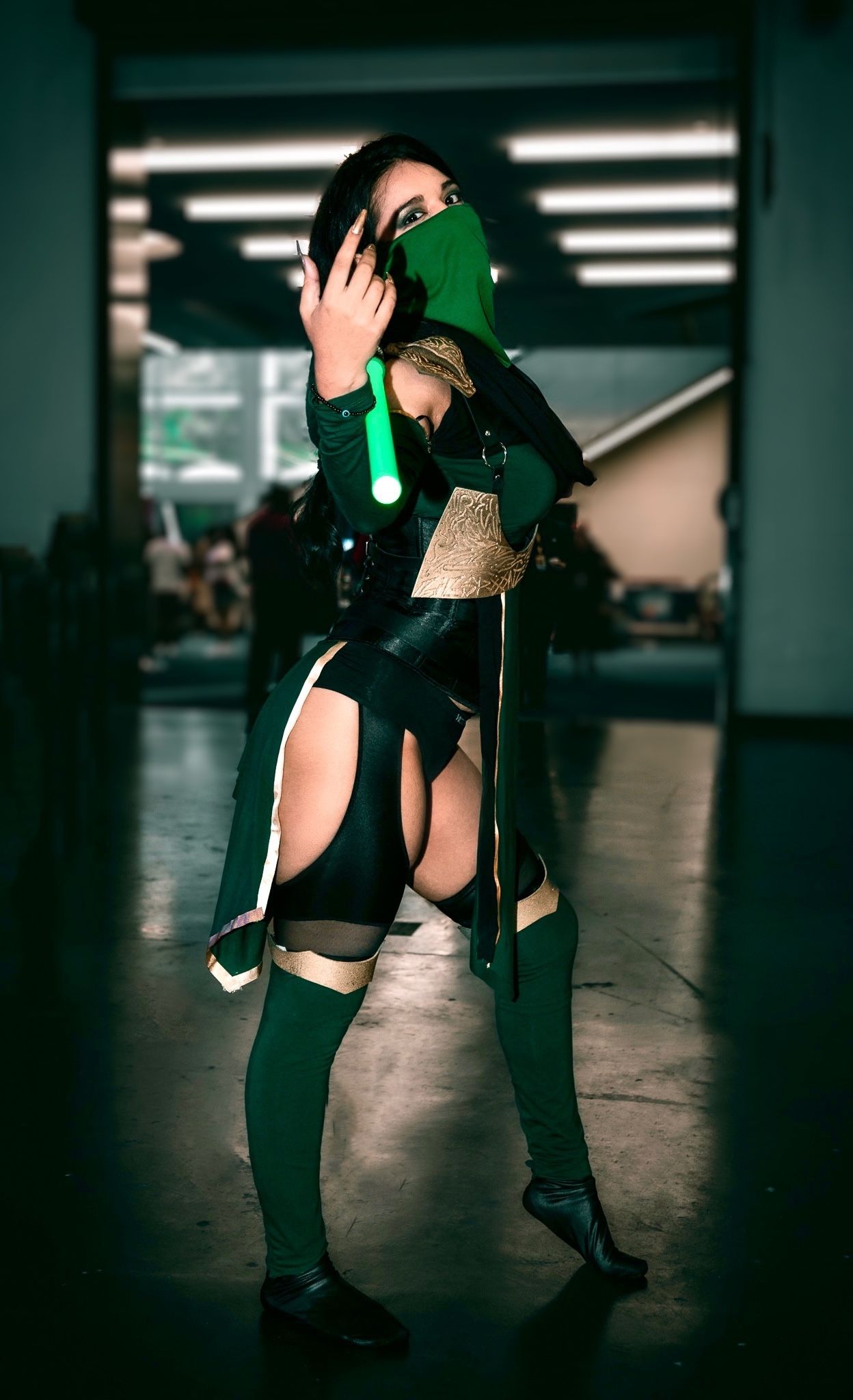 Cosplayer in dark green and black outfit poses dramatically in dimly lit convention space.