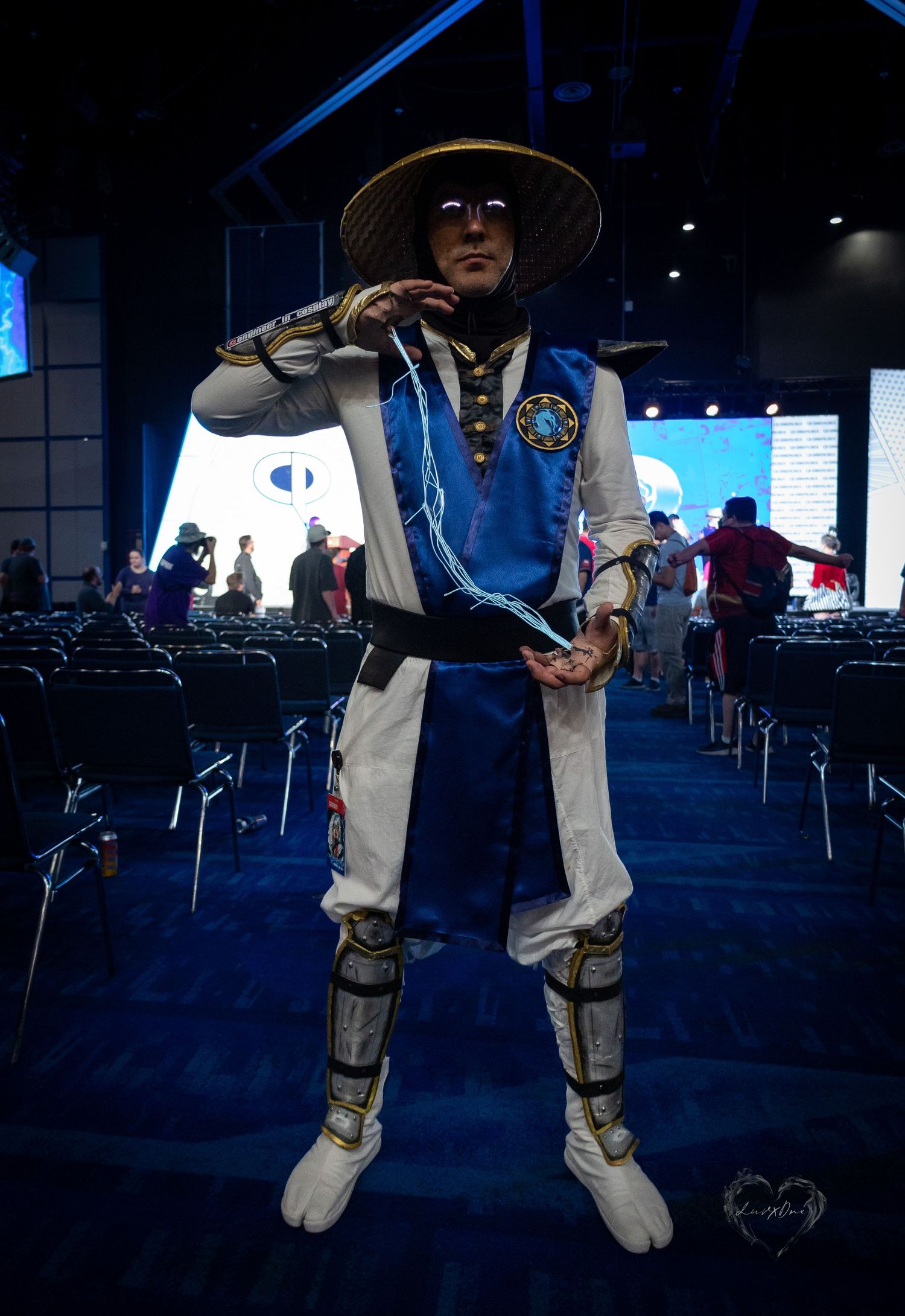 Cosplayer in Mortal Kombat inspired blue and white outfit with conical hat at gaming convention.