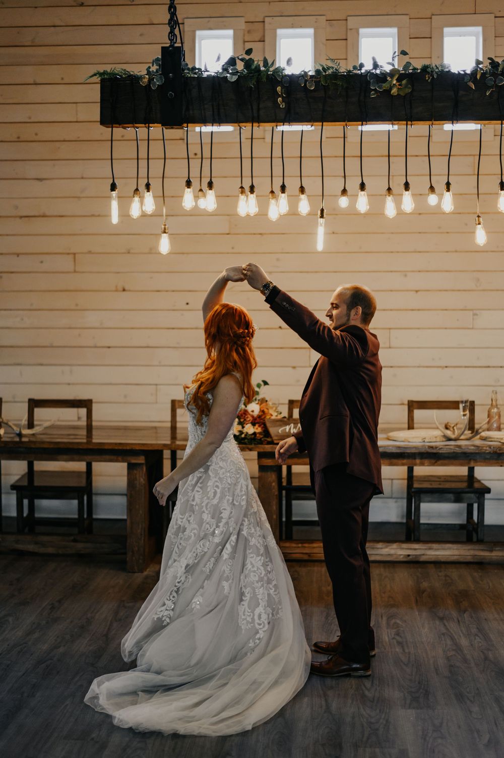 A wedding couple dances under hanging Edison bulbs in a rustic wooden venue.