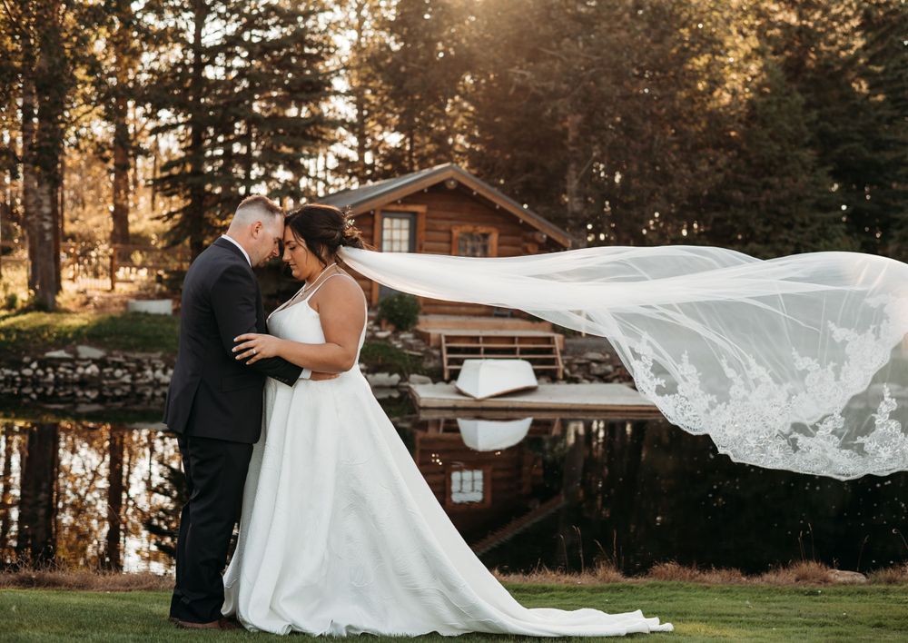 A dramatic wedding portrait by a lake with flowing veil and rustic cabin in the background.