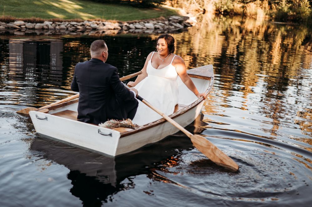 A wedding couple sits in a white rowboat on a tranquil lake during golden hour with cliffs in the background.