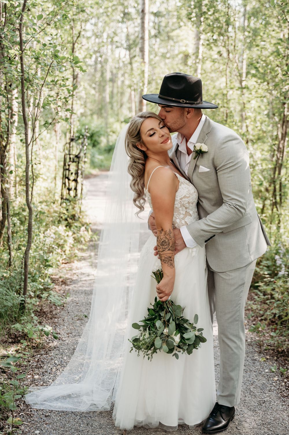 Newlyweds share an intimate moment on a forest path with sunlight filtering through the trees.