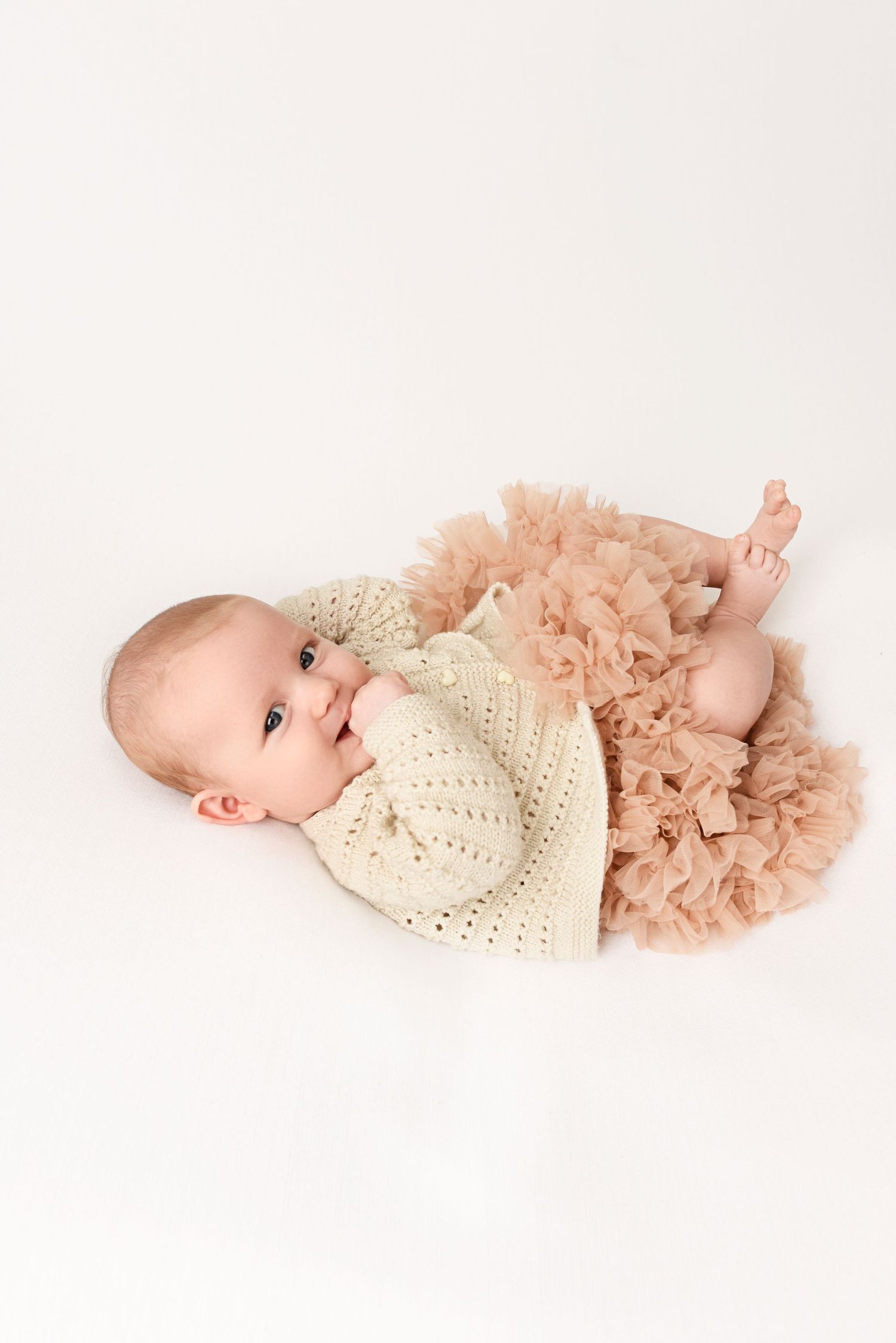 A baby wearing cream knit sweater and peach tulle skirt poses on white background.