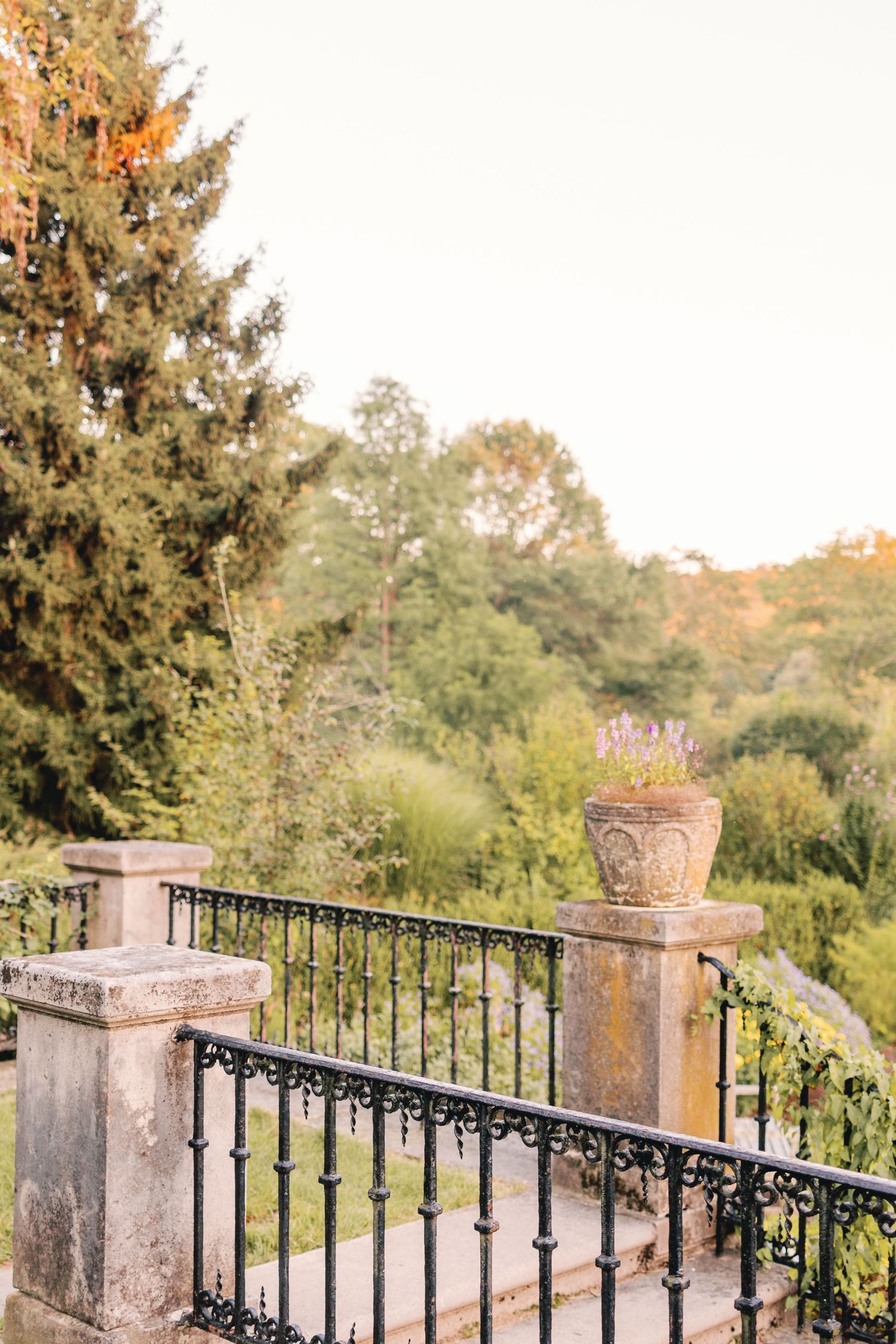 Stone balcony with ornate iron railing and decorative urn overlooking lush garden at sunset.
