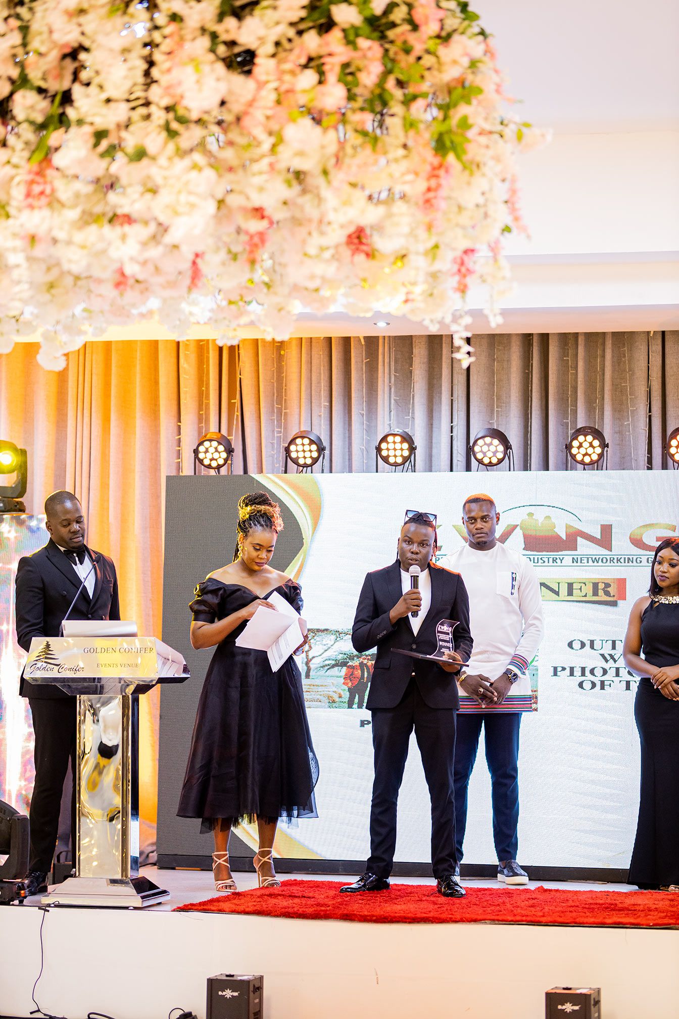 Corporate event presentation with speakers on stage under floral decor and dramatic lighting.