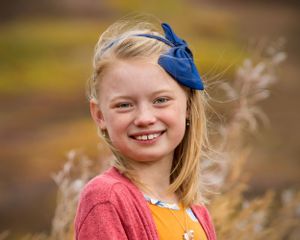 Young person wearing coral sweater and blue bow poses outdoors during autumn.