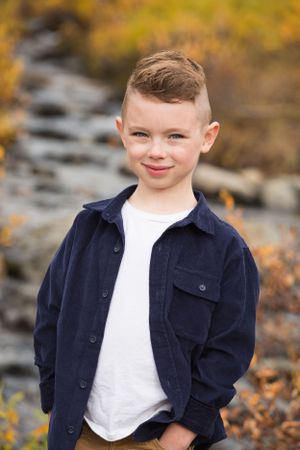 Young person in navy jacket and white shirt poses outdoors in autumn setting.