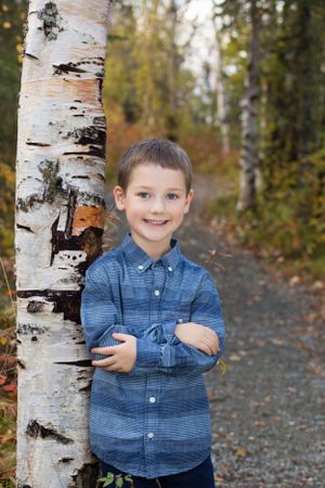 Young person in blue button-down shirt poses beside birch trees on autumn trail.