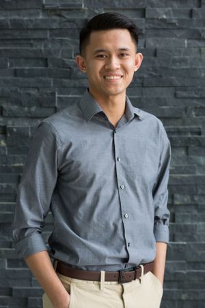 Young person in grey dress shirt and khakis poses against stone wall background.