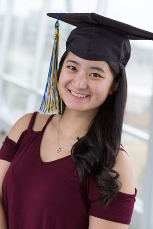 Graduate in burgundy dress and black cap smiles during indoor photoshoot.