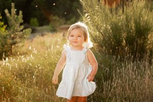 Child in white dress stands among tall grasses during golden hour sunset.