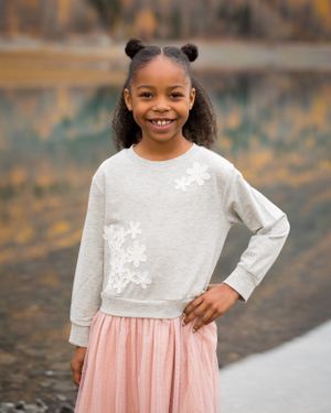 Child wearing white sweater and pink skirt poses confidently against autumn mountain backdrop.