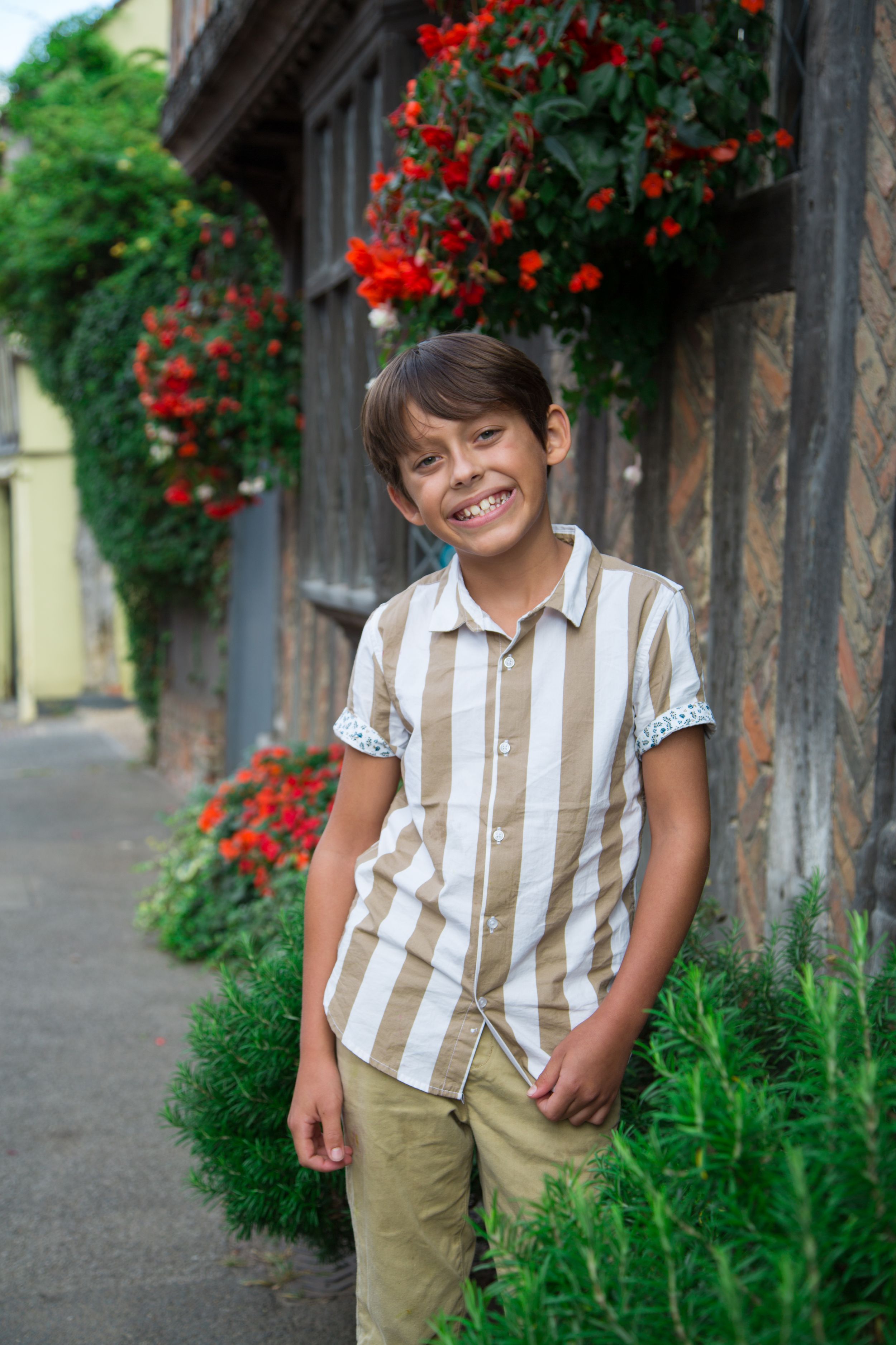 Young person in striped shirt and khakis poses near red flowers and wooden columns outdoors.