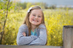 Young person smiling while leaning on wooden fence post surrounded by bright yellow wildflowers in springtime.