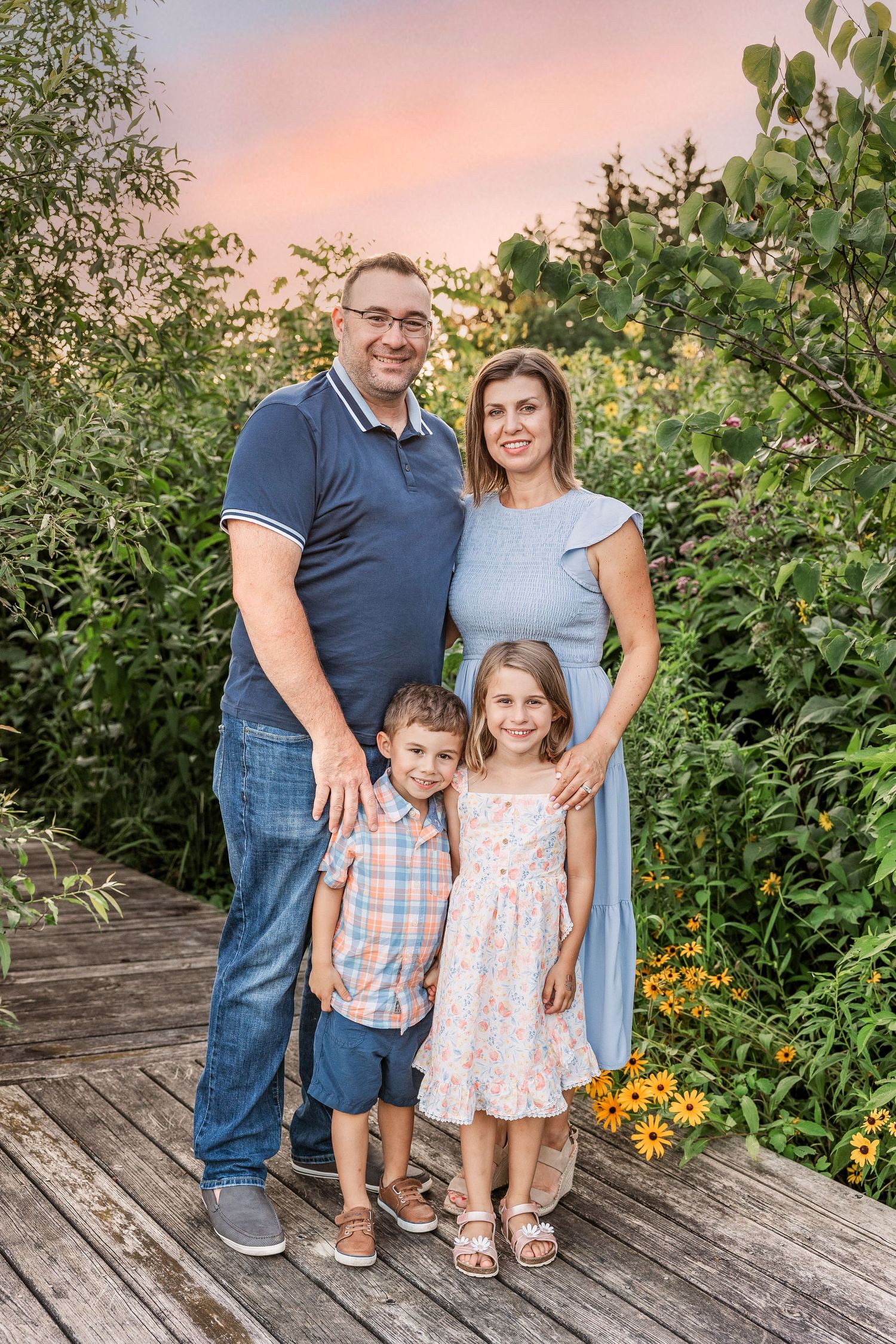 Family of four poses together on wooden deck with sunset and greenery in background.