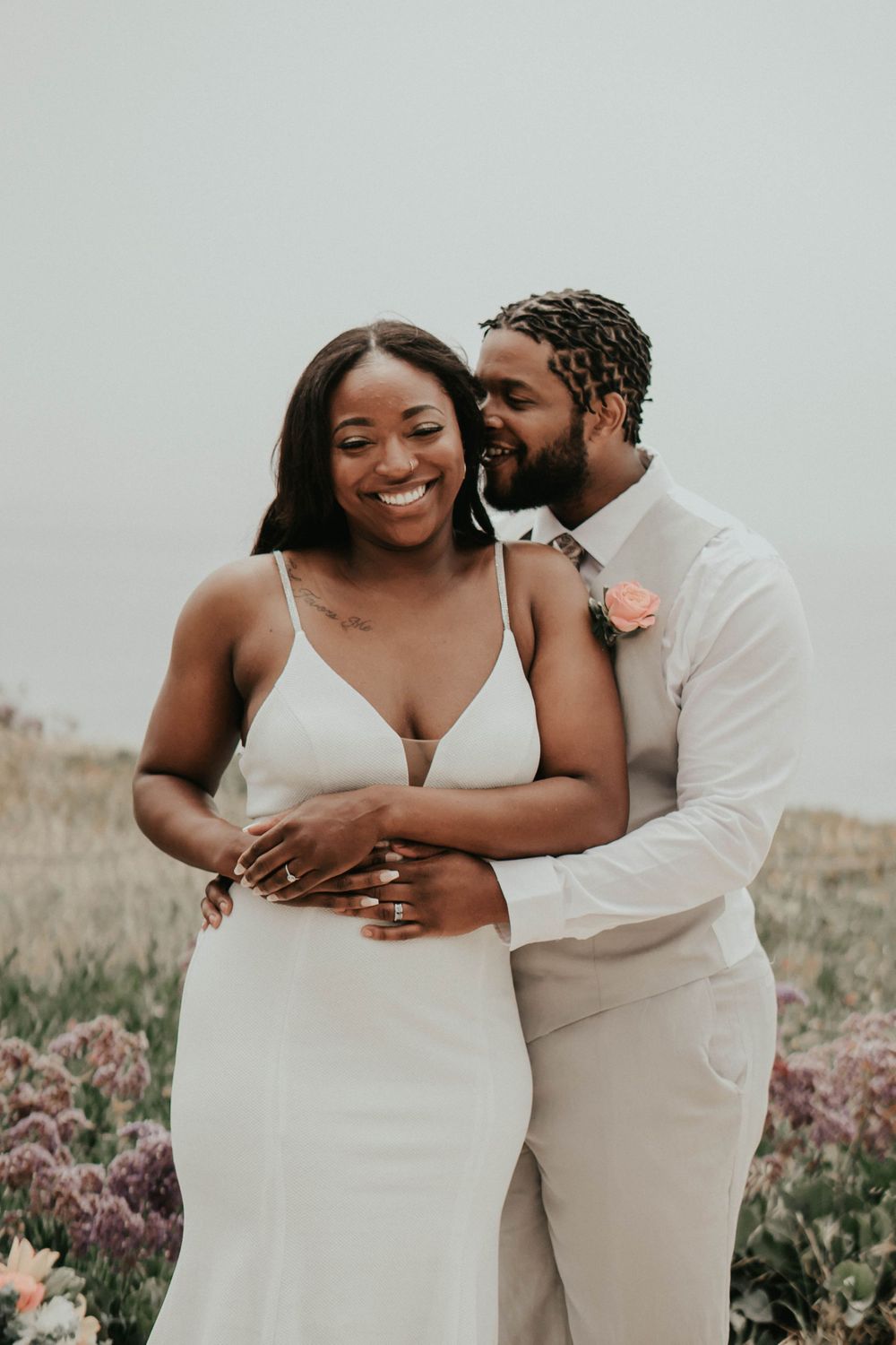 Newlywed couple embracing in white attire against natural outdoor setting.