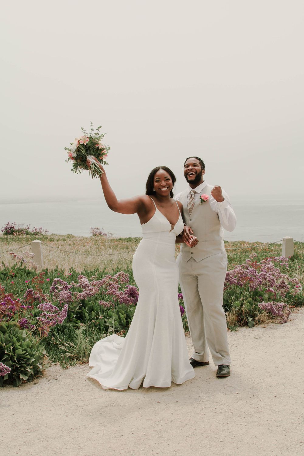 Wedding couple celebrating with bouquet raised against coastal backdrop.