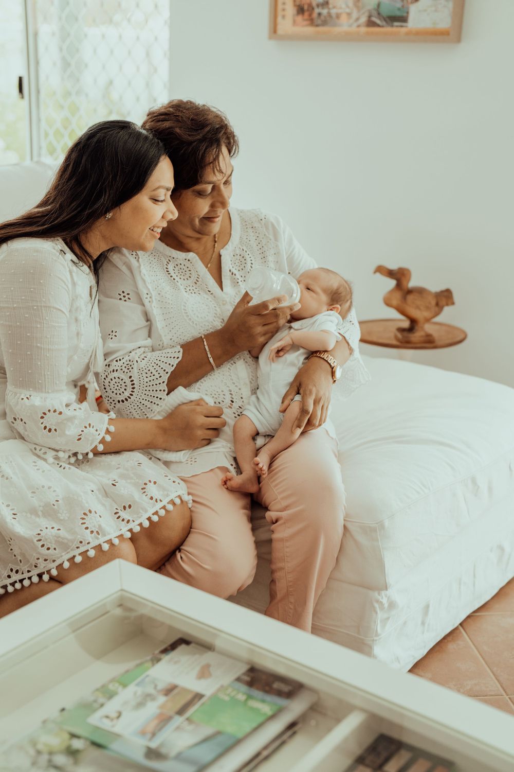 Two people in white lace dresses holding infant on white couch.