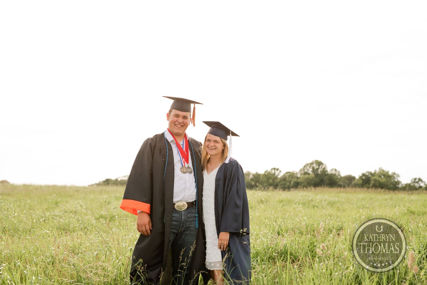 Two graduates in caps and gowns pose together in a sunny field at sunset.