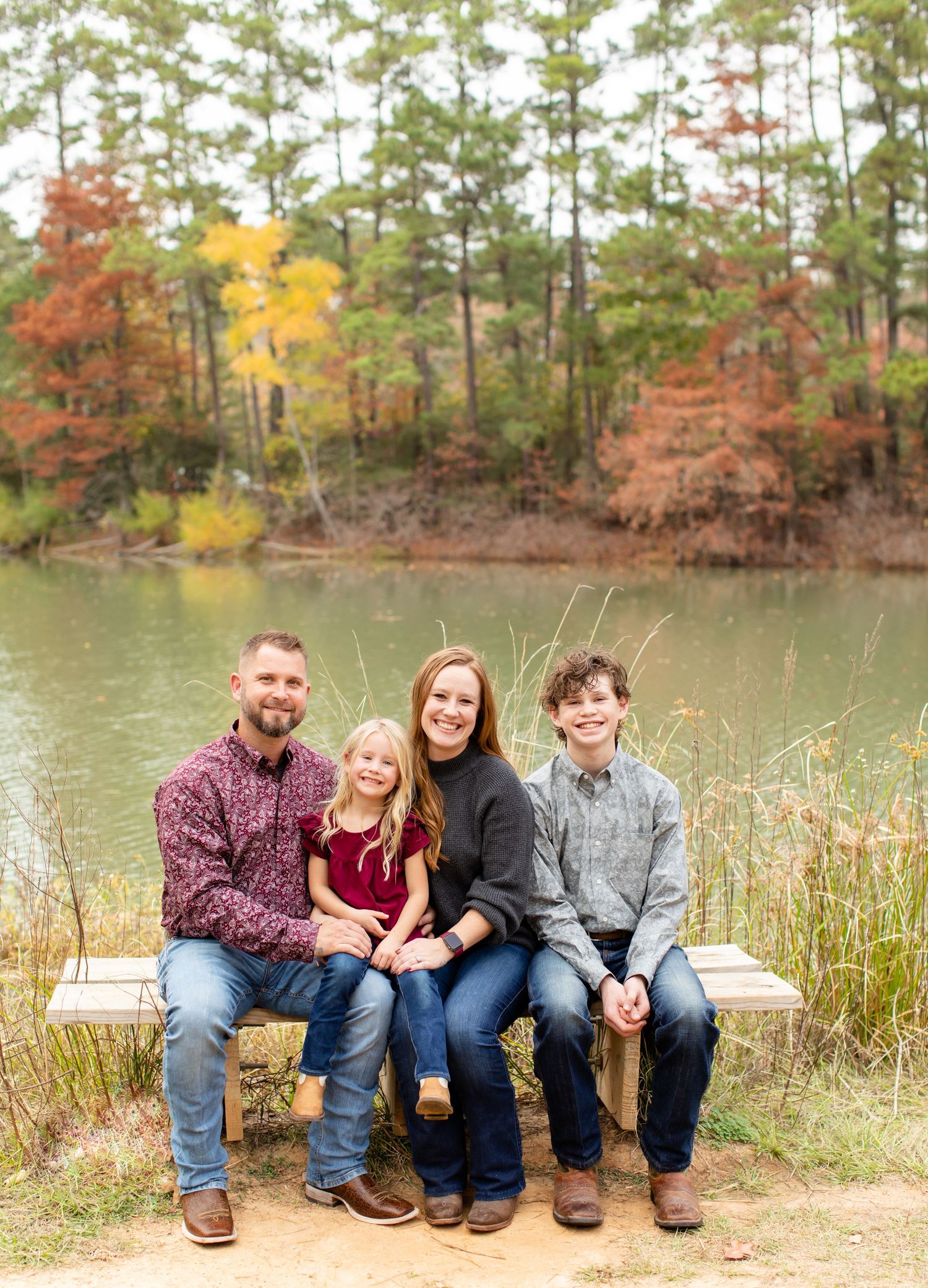 A family sits together on a wooden dock overlooking a lake surrounded by autumn trees.