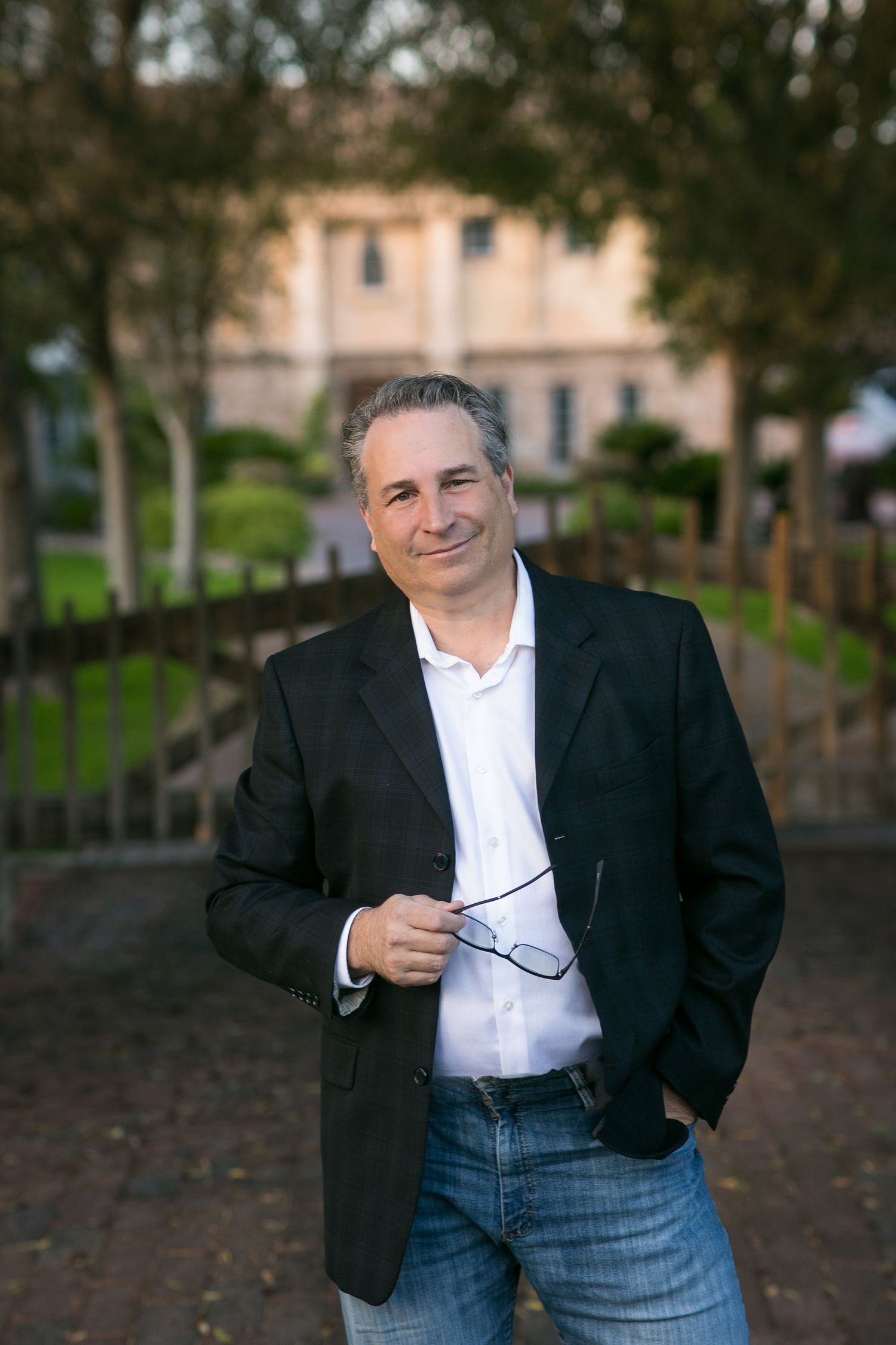 Smiling middle-aged man in black blazer holding glasses outdoors near park with blurred building background.