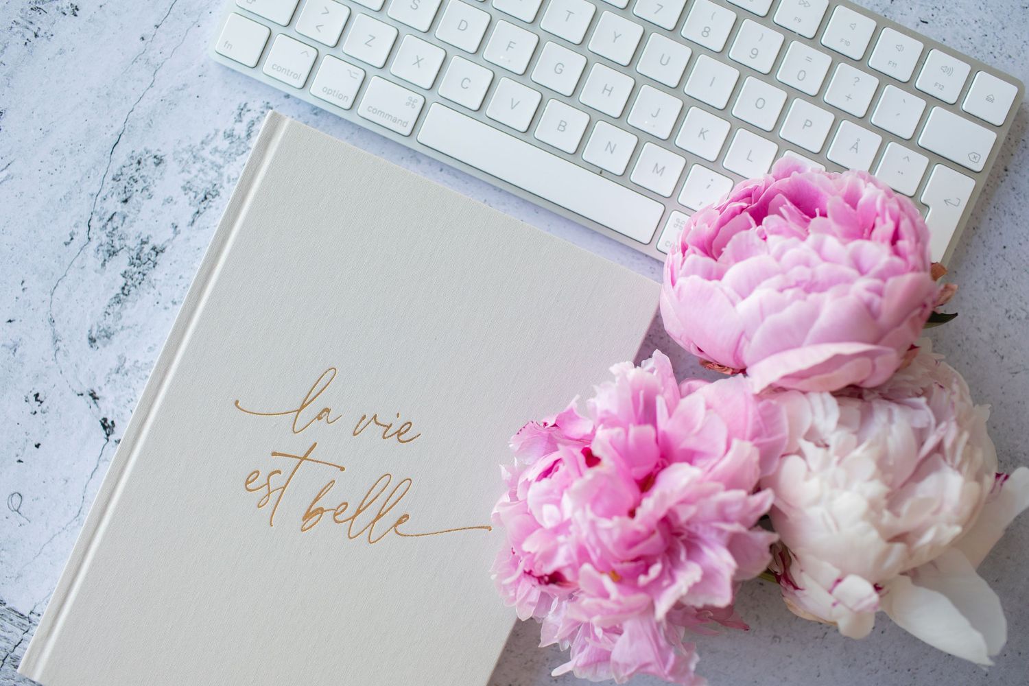 Pink peonies rest beside a white keyboard and notebook with gold text on a marble surface.