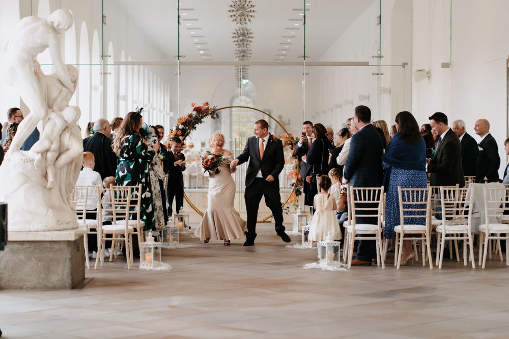 Wedding ceremony in modern white venue with gold arch and white chairs at The Orangery Margam Park