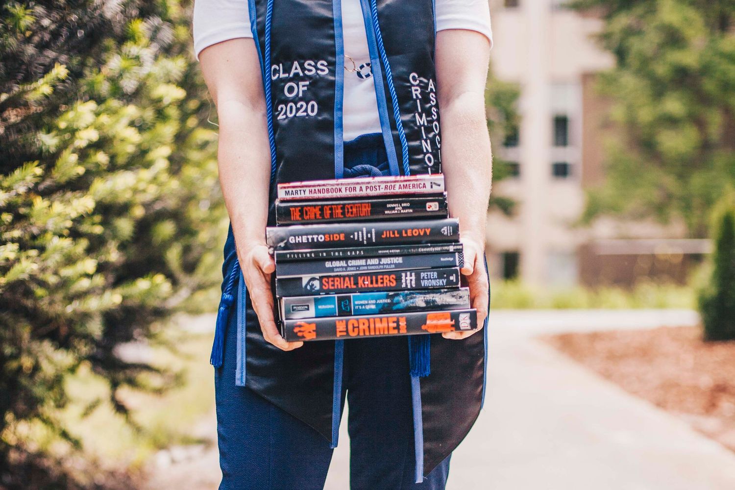 Graduate holding a tall stack of crime and legal themed books while wearing a Class of 2020 stole.